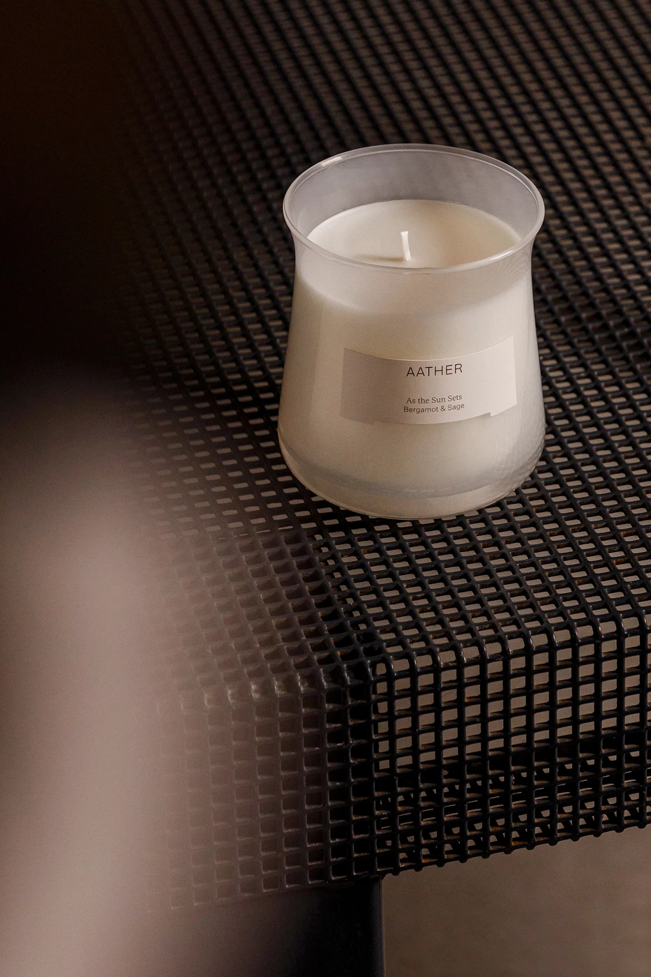 Aather scented candle in a frosted glass jar resting on a dark grid surface with a soft blur in the foreground.