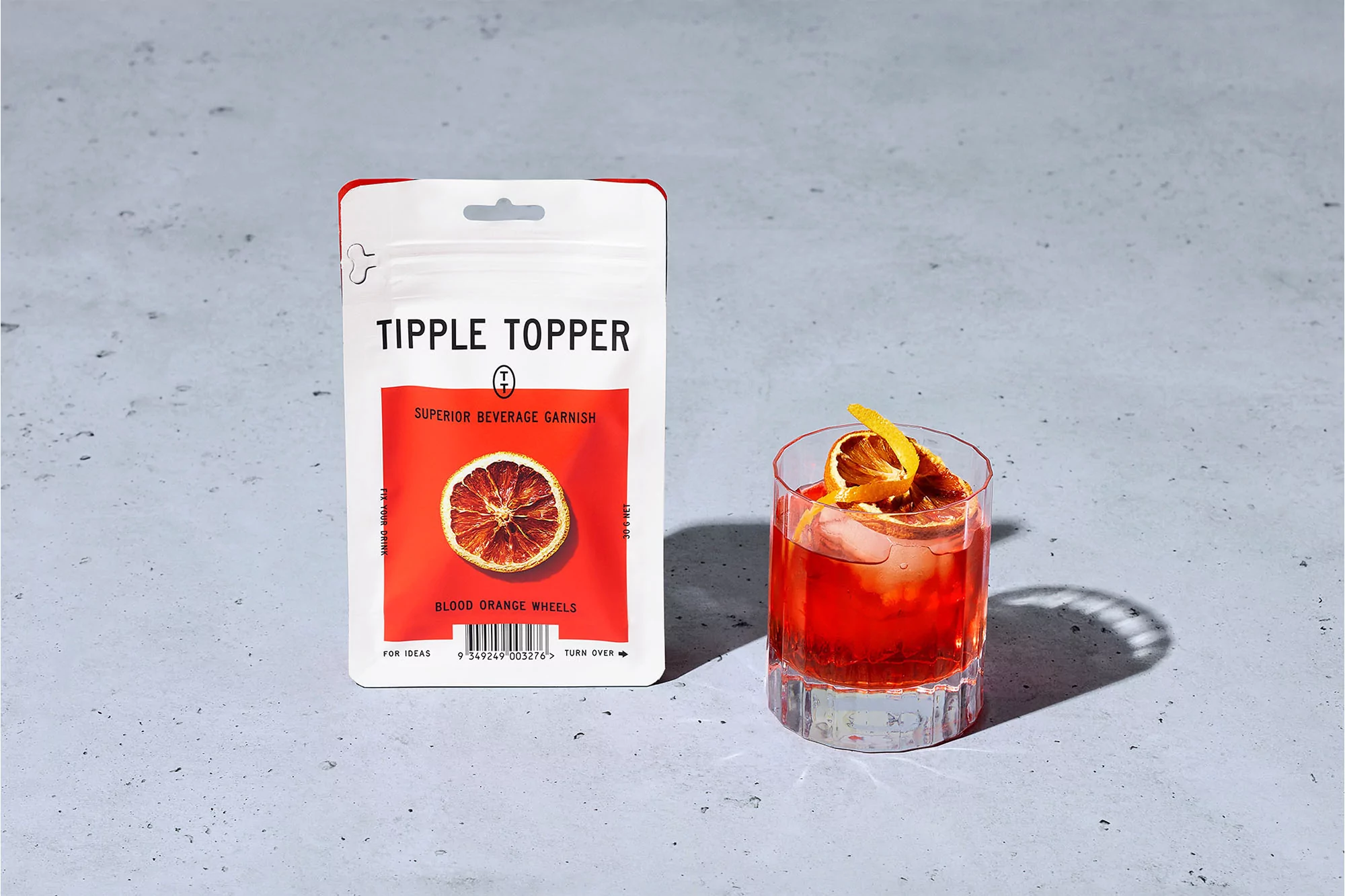 Cocktail in a glass with blood orange garnish beside a package of Tipple Topper dried citrus slices on a concrete surface.