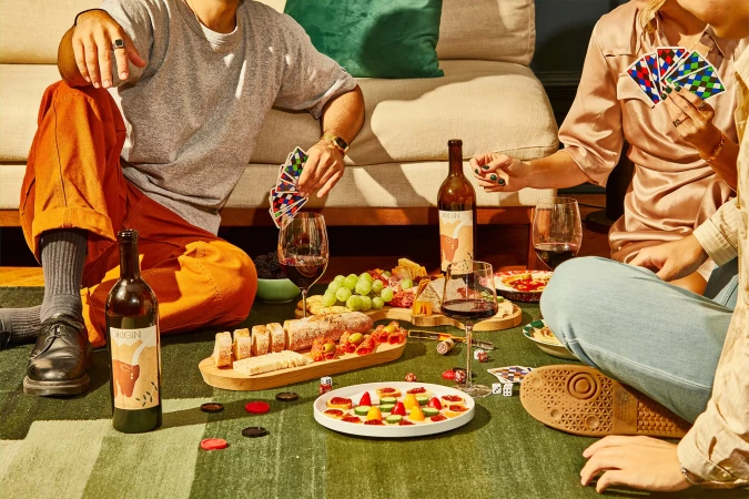 Three people sit on a green rug playing cards surrounded by wine, snacks, and playing chips in a relaxed indoor setting with warm lighting.