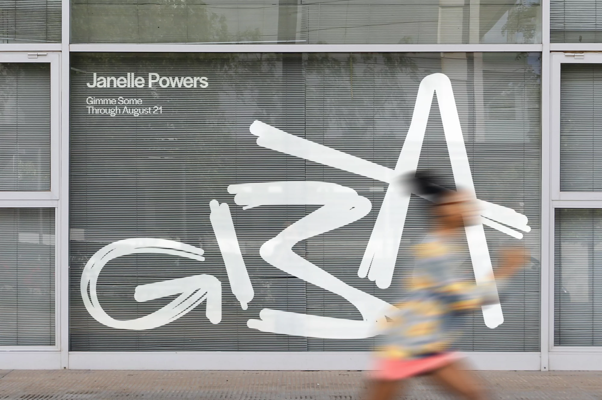A blurry photo of a person walking past a building with a large white graphic on the window, advertising Janelle Powers' art.