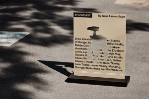 A freestanding sign with cutout typography and black lettering stands on asphalt under mottled tree shade.