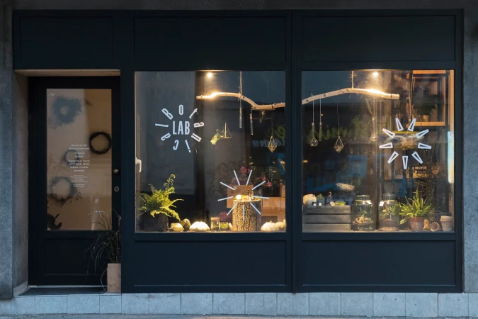 A storefront window displays terrariums and plants in a dark-framed structure on a city street, viewed from a slightly low angle.