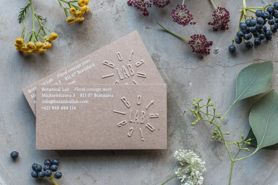 Botanical Lab business cards with floral accents on concrete backdrop, featuring yellow tansy, dark berries, and fresh eucalyptus.