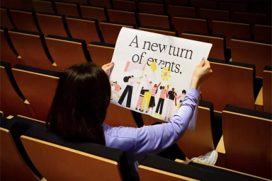 Woman holding open printed newspaper with graphic illustration in empty auditorium with rows of seats