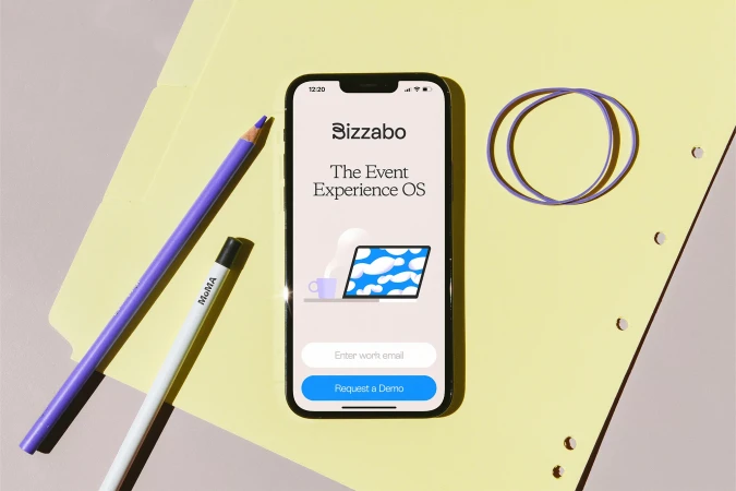 A smartphone displaying the Bizzabo app on a yellow folder beside purple pencils and rubber bands under bright lighting.