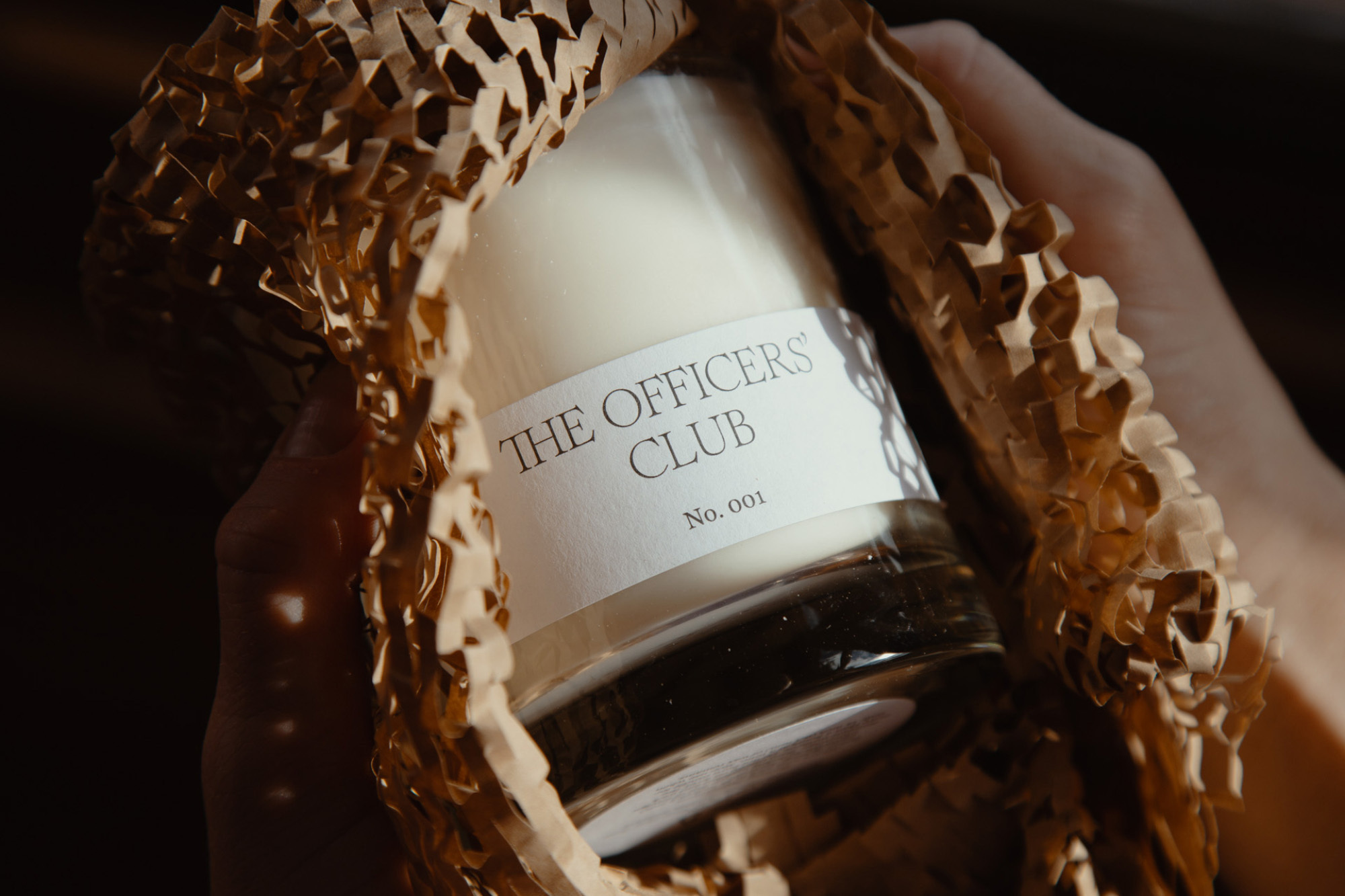 Close-up shot of a clear glass jar with white candle wax inside, held by two hands and packaged in shredded brown paper.