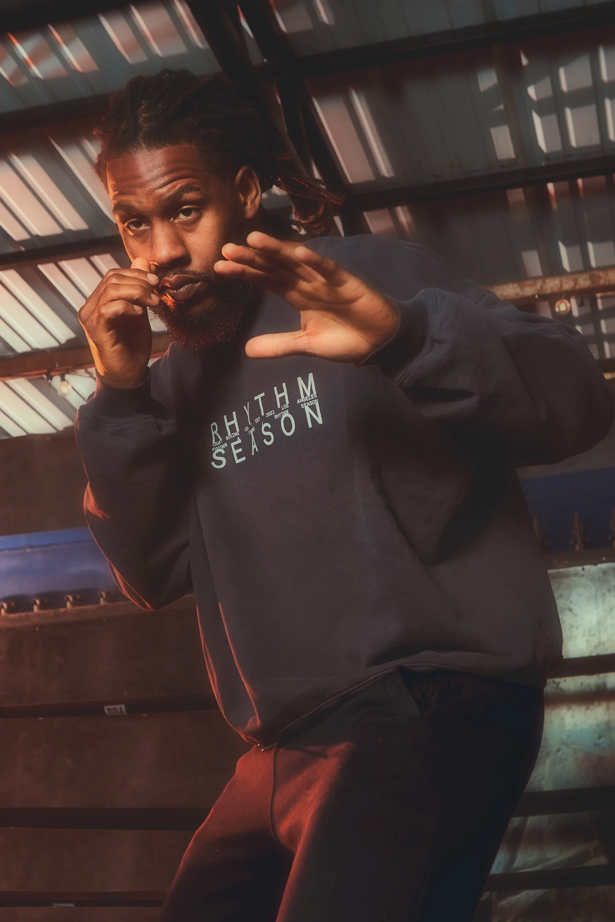 An adult Black man in a sweatshirt raises his hands in a boxing stance in an indoor setting with a metal structure above.