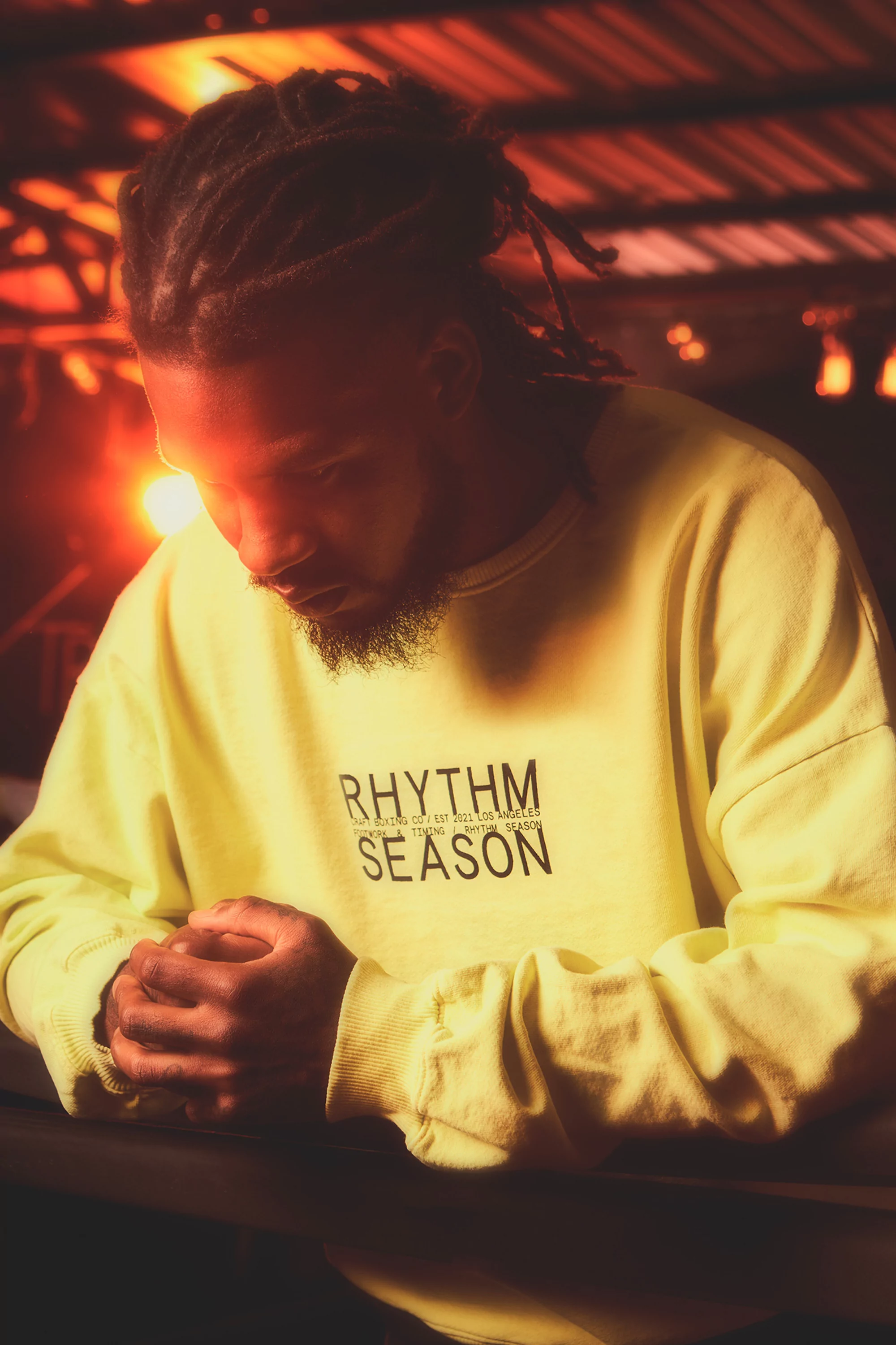 Portrait of a man with dreadlocks in a yellow sweatshirt. He is indoors with warm lighting and a dark background. His hands are clasped.