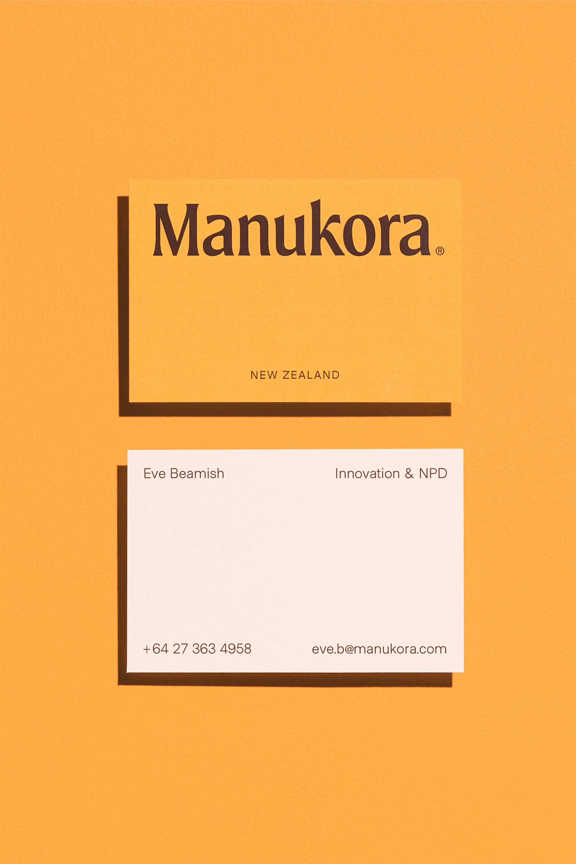 Two business cards, one cream with the Manukora logo, one white with contact information, on a light yellow background.