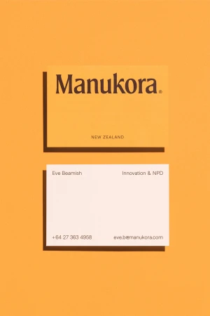Two business cards, one cream with the Manukora logo, one white with contact information, on a light yellow background.