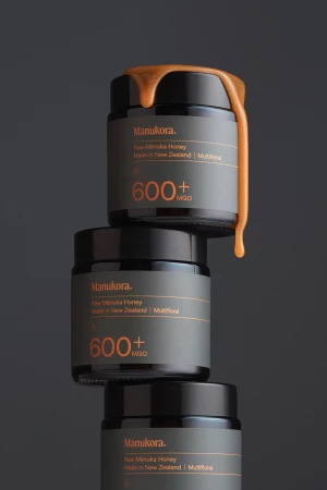 Tower of Manukora raw Manuka honey jars with gold honey dripping, against a dark gray studio backdrop, product photography.