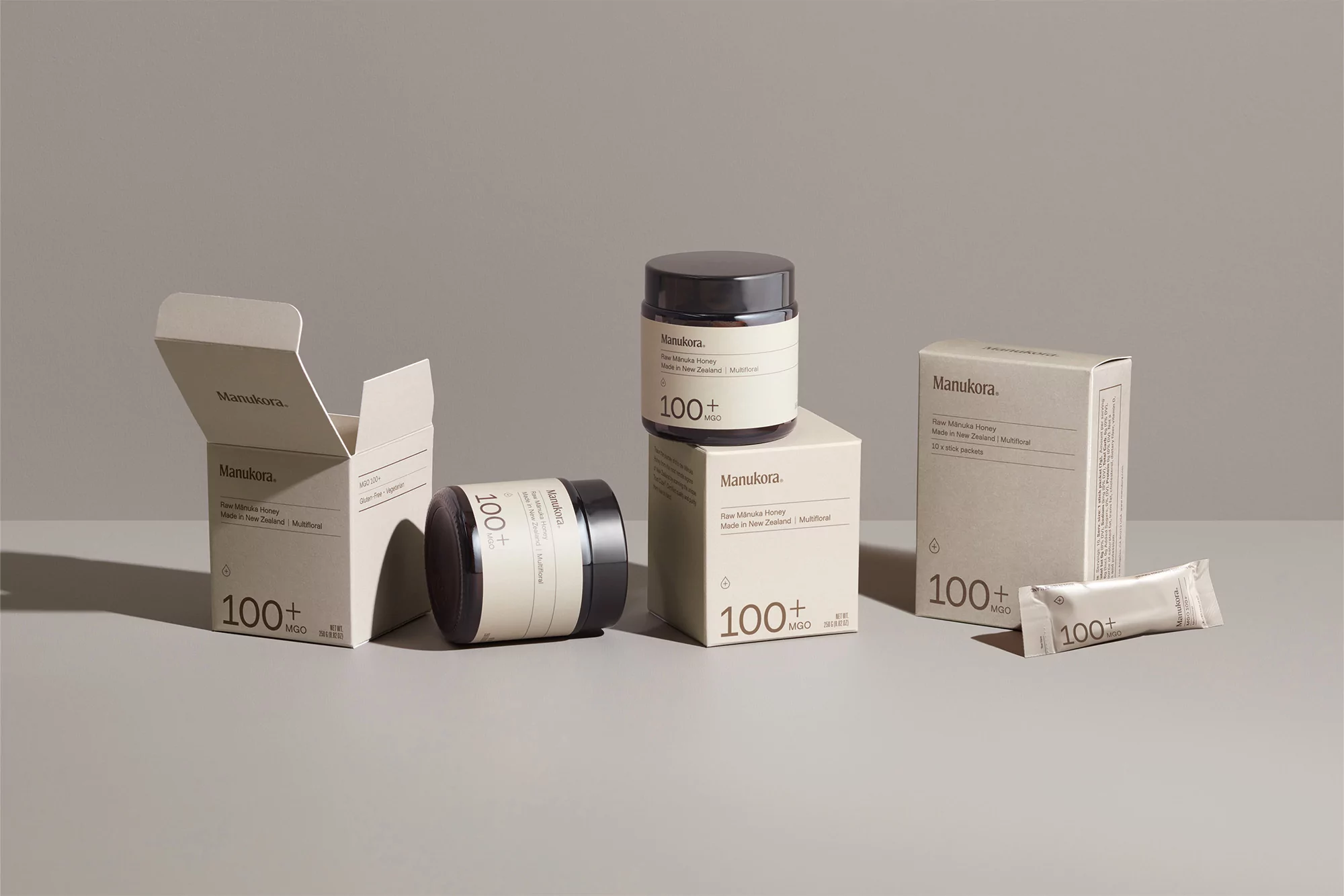 Several jars and boxes of Manukora honey products are displayed on a gray surface, showcasing their packaging and branding details.