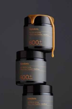 Three stacked dark honey jars with golden raw manuka honey dripping from the top rim against a dark grey background.