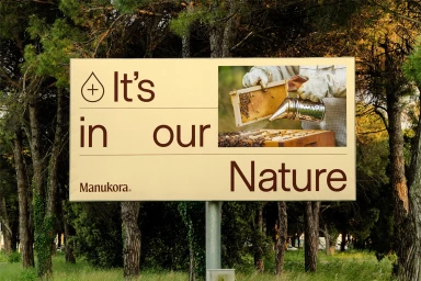 Manukora brand identity billboard displaying text and a beekeeper image outdoors among tall trees