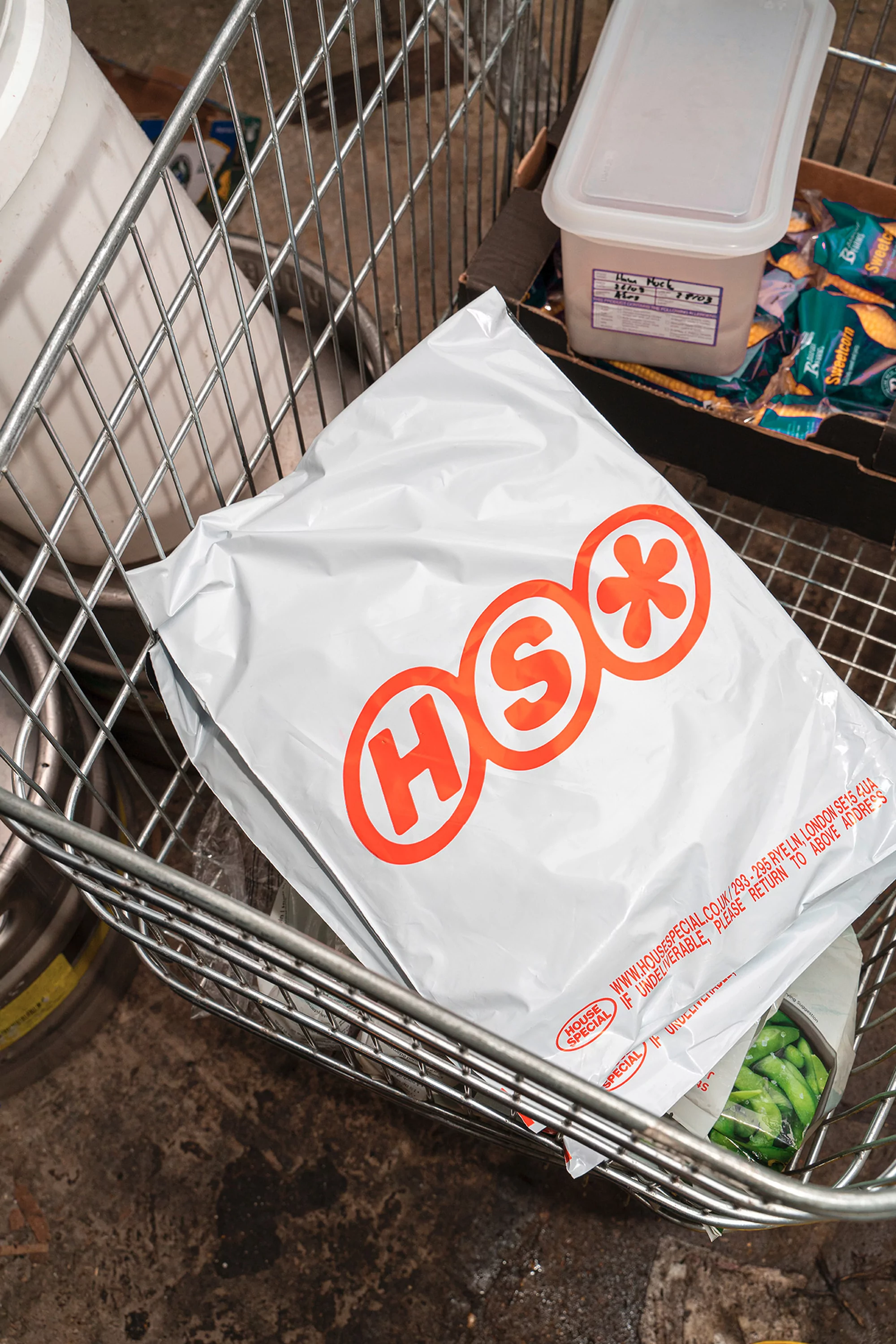 Package with 'HSO' logo in a wire shopping cart, surrounded by a storage container, packaged food and a dark concrete floor.