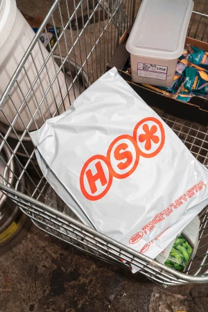 Package with 'HSO' logo in a wire shopping cart, surrounded by a storage container, packaged food and a dark concrete floor.