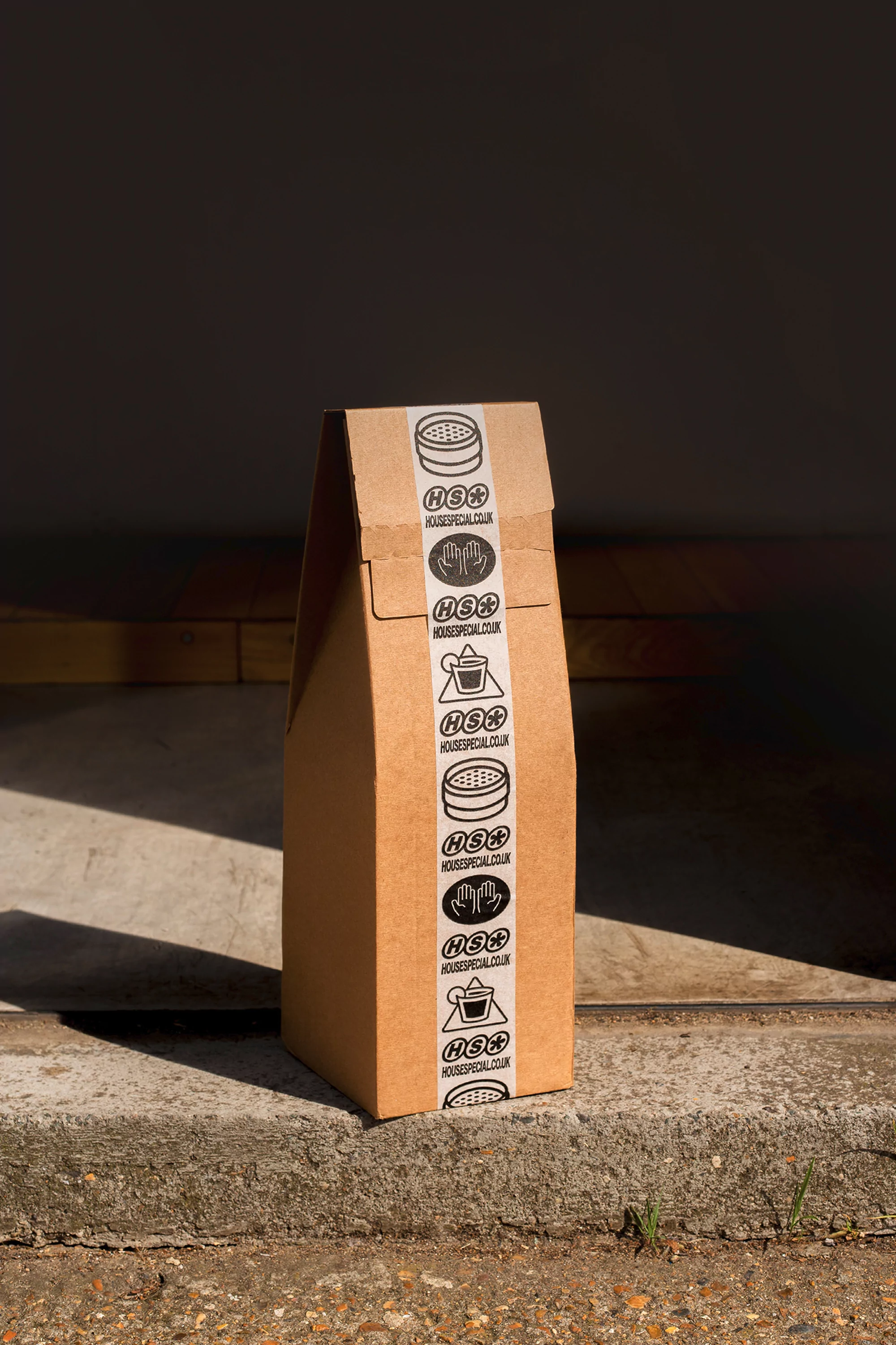 Brown paper bag with logo on concrete surface against dark background, casting light and shadow with natural textures.