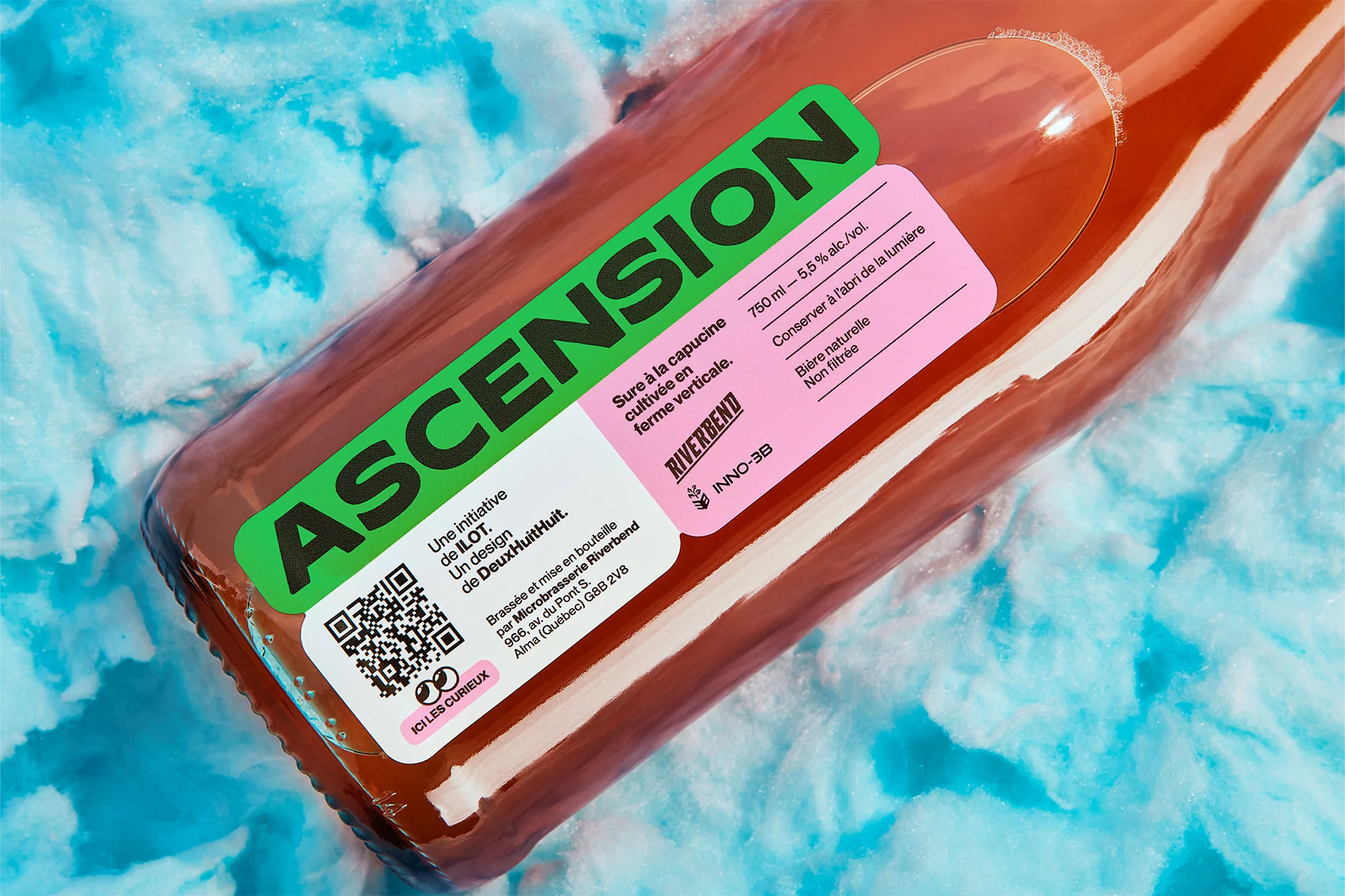 Close-up of a bottle of Ascension beer with a label featuring a QR code and a pink section with the beer's details against a blue backdrop.