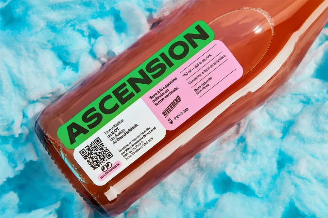 Close-up of a bottle of Ascension beer with a label featuring a QR code and a pink section with the beer's details against a blue backdrop.