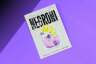 A white rectangular print featuring an illustration of a cocktail and bold typography, resting on a textured purple surface.