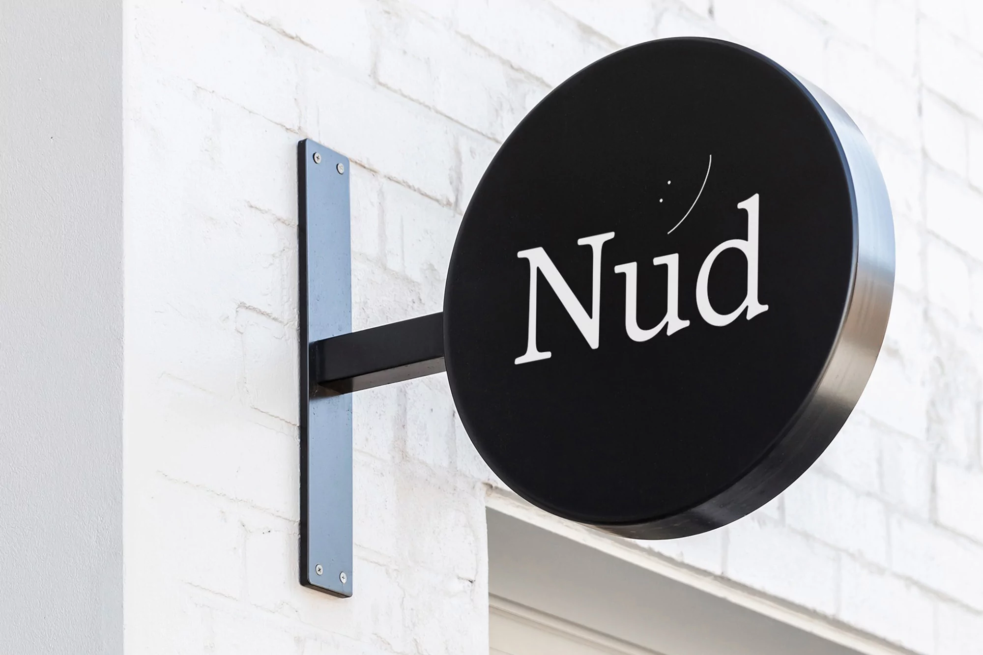 Outdoor sign for "Nud" is mounted on a white brick wall with black metal hardware. The black circular sign features white lettering.