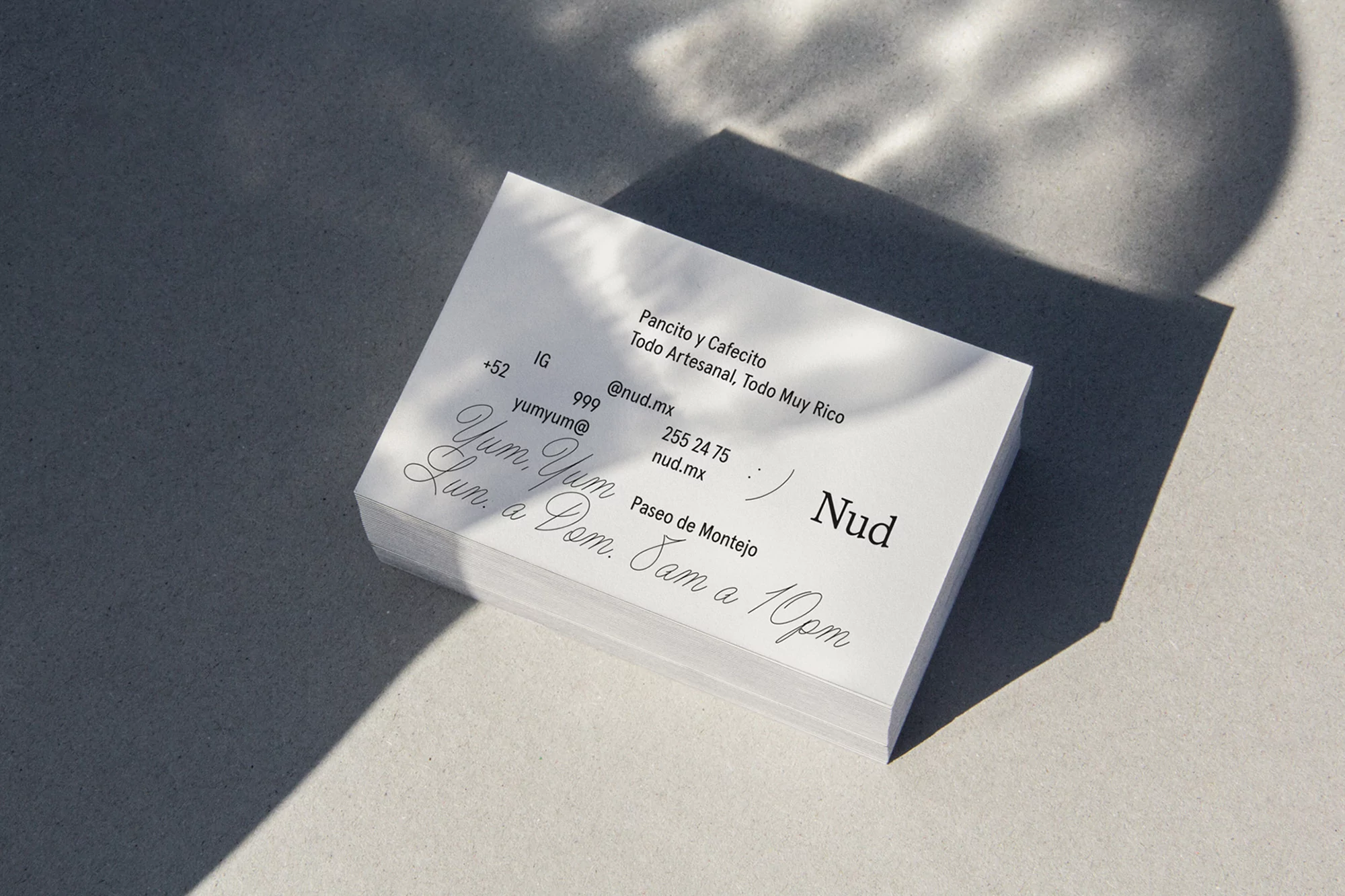 Stack of white business cards on a neutral surface with sunlight and shadows, featuring the logo and contact information for 'Nud'.