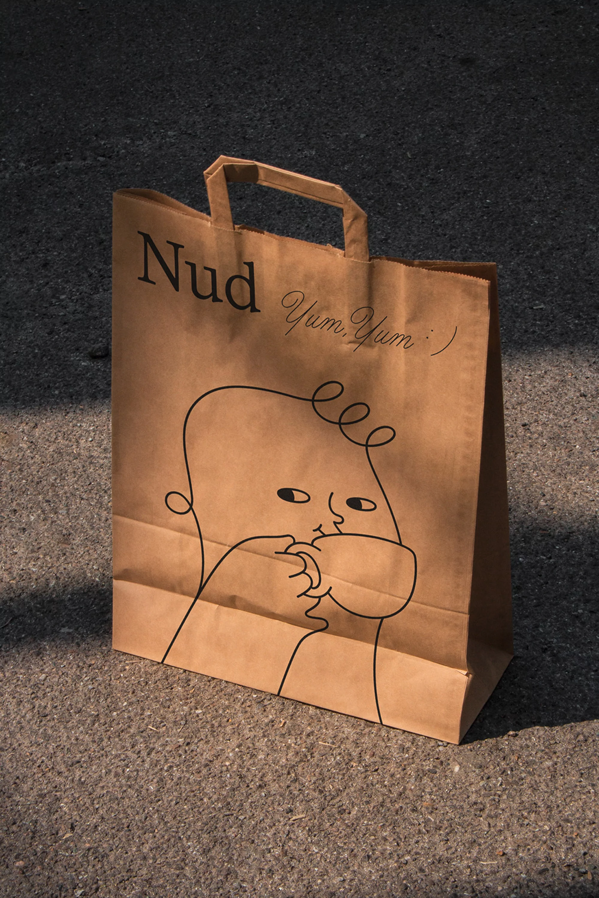 Close-up of a brown paper shopping bag with a whimsical line drawing of a child drinking and the text 'Nud Yum Yum' on a grey surface.