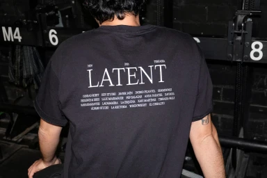 Person seen from behind wearing a black t-shirt printed with LATENT branding and various designer names.