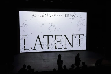 Silhouettes of people walking past a large white screen displaying graphic typography and the word LATENT in a dark venue.