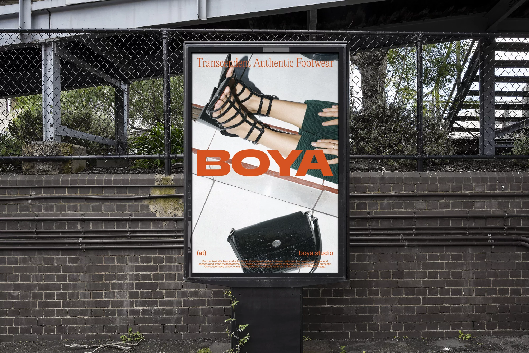 Outdoor billboard displaying a fashion advertisement featuring black strappy heels, green fabric, and a black handbag against a brick wall.