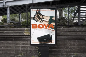 An outdoor advertising poster display for The RaDD featuring strapped sandals and a black handbag mounted on a brick wall.