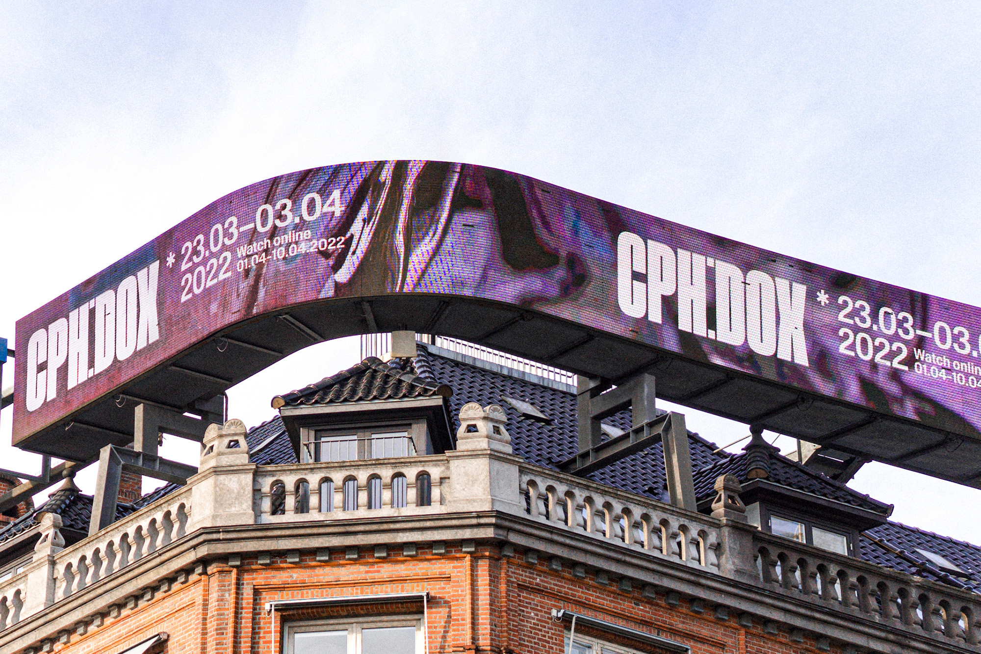A large CPH:DOX billboard advertisement is mounted on top of a brick building against a bright, overcast sky.