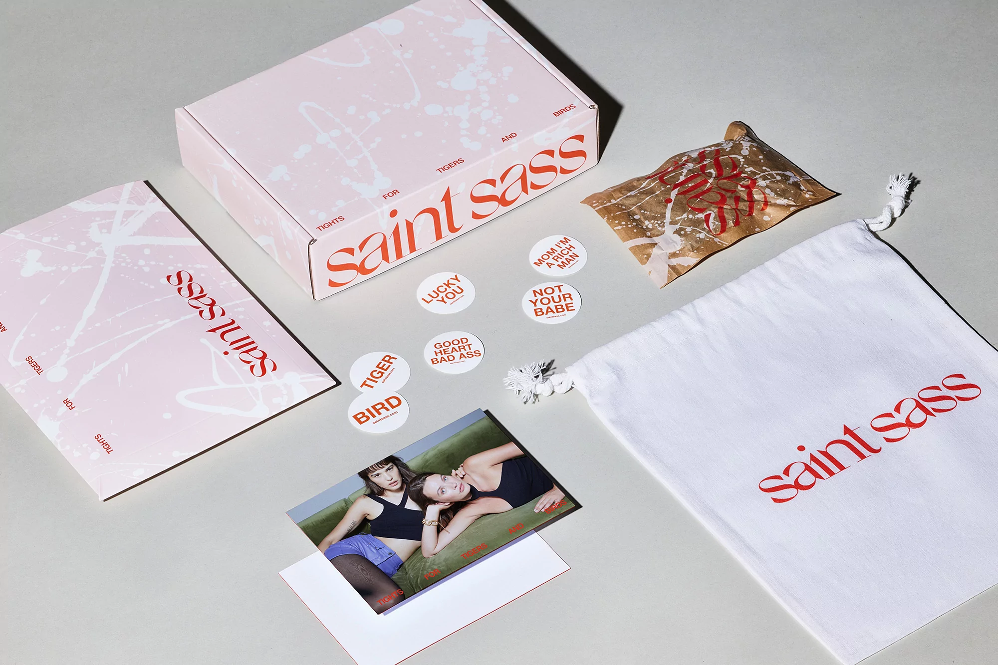 A flat lay of Saint Sass brand items including packaging, round stickers with text, and a photo print on a neutral background.