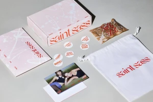 Product packaging flat lay for Saint Sass featuring a pink branded box, white dust bag, stickers, and promotional photo.