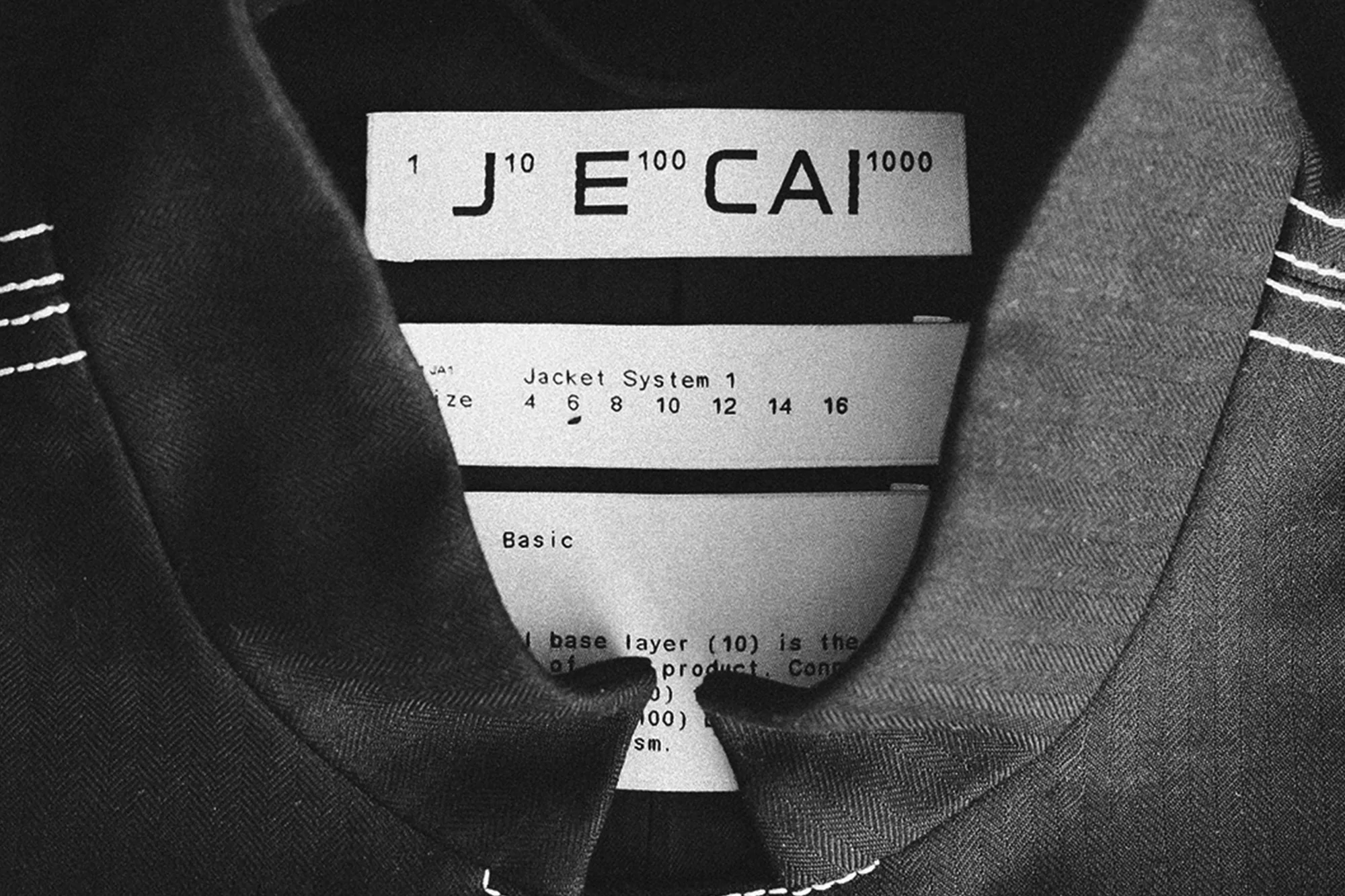 Close-up of a garment tag with the brand name J'E CAI and jacket sizing information stitched onto a dark grey jacket.