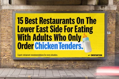 Yellow billboard with black text and baby bottle graphic mounted on a brick wall above a sidewalk.