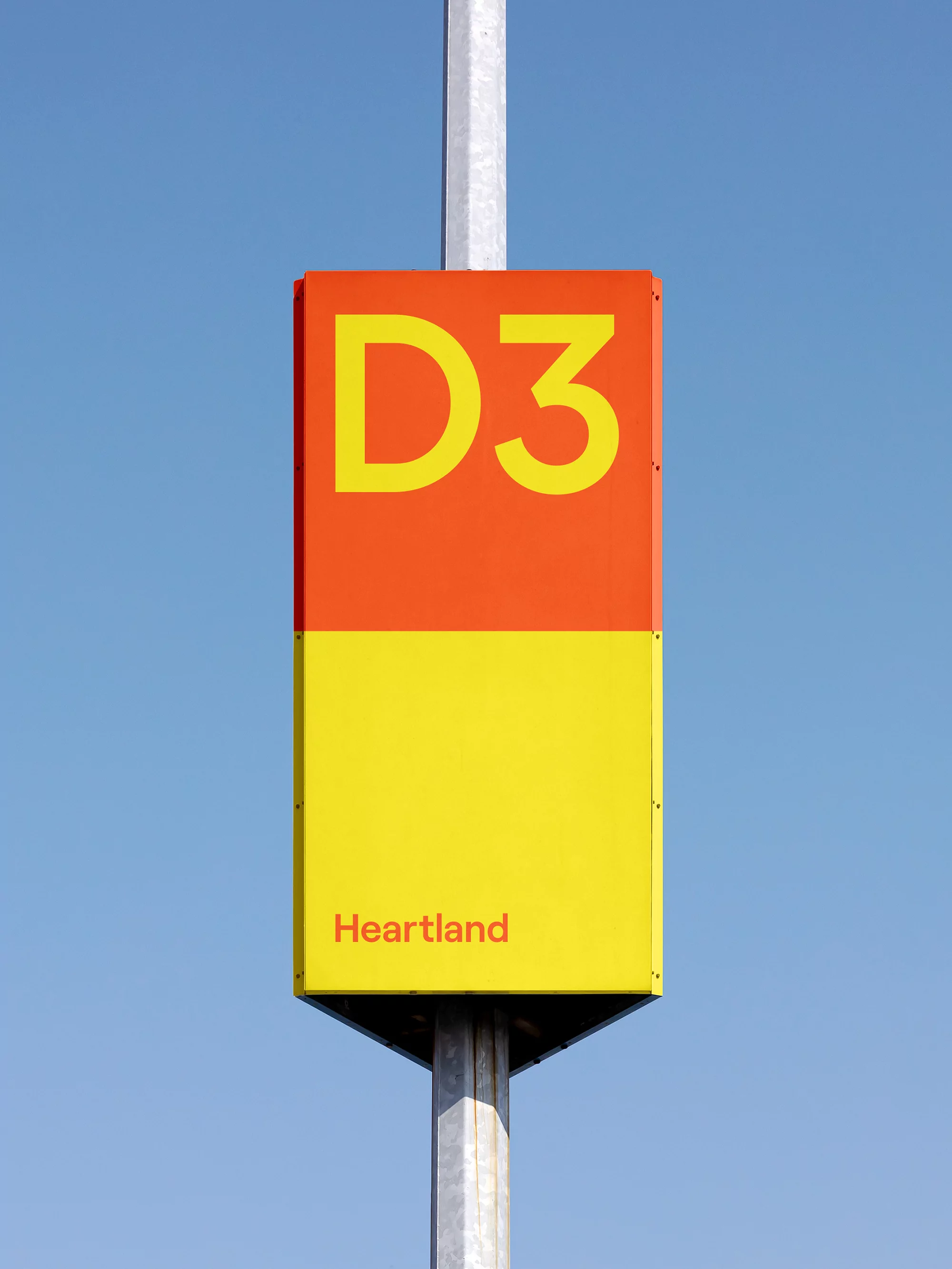 Orange and yellow rectangular sign with 'D3' and 'Heartland' on a gray metal post against a clear blue sky in an outdoor setting.