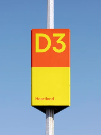 Orange and yellow rectangular sign with 'D3' and 'Heartland' on a gray metal post against a clear blue sky in an outdoor setting.