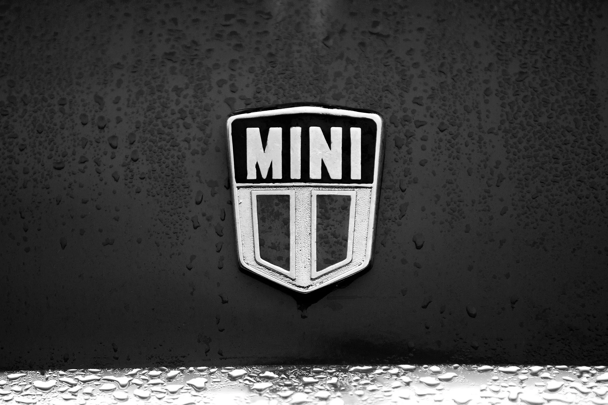 Close-up of a black car hood featuring a silver MINI logo emblem with water droplets reflecting light, creating a textured surface.