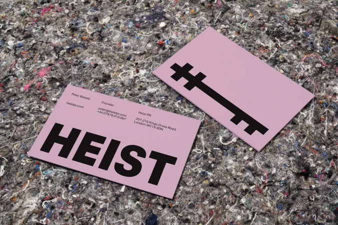 Two pink business cards with black text and graphics on a textured grey surface, one angled to show a logo.