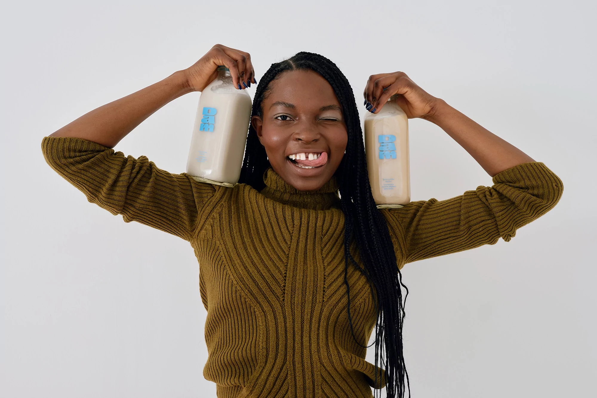 Smiling woman holds two bottles of beverage over her shoulders indoors against a white wall, sticking her tongue out.