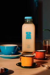 Glass bottle of dairy beverage with light blue cap and DAM logo surrounded by colourful cups on a wooden table.