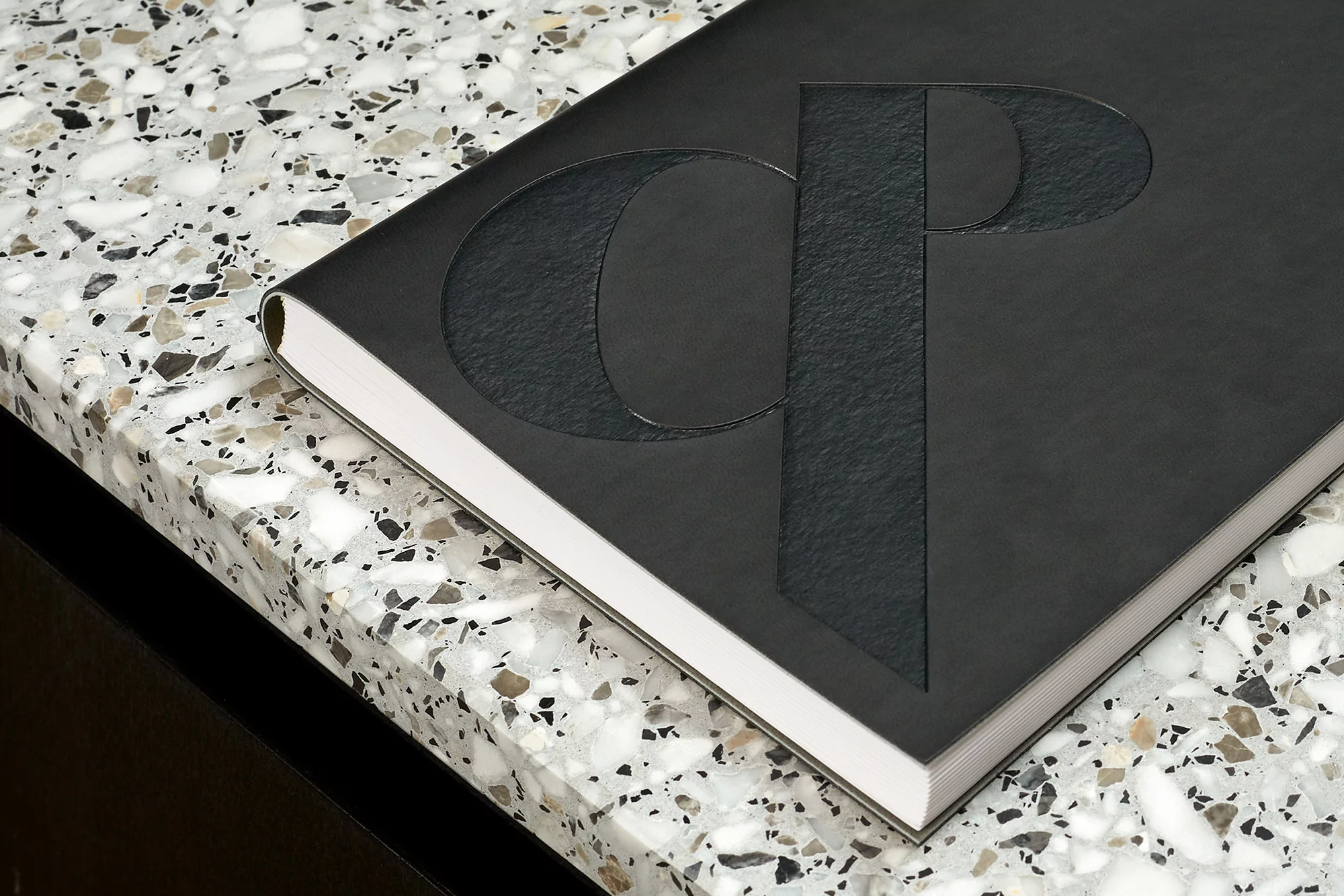 Close-up of a black hardcover book with a textured embossed CP monogram on a speckled surface, emphasizing material and minimalist design.