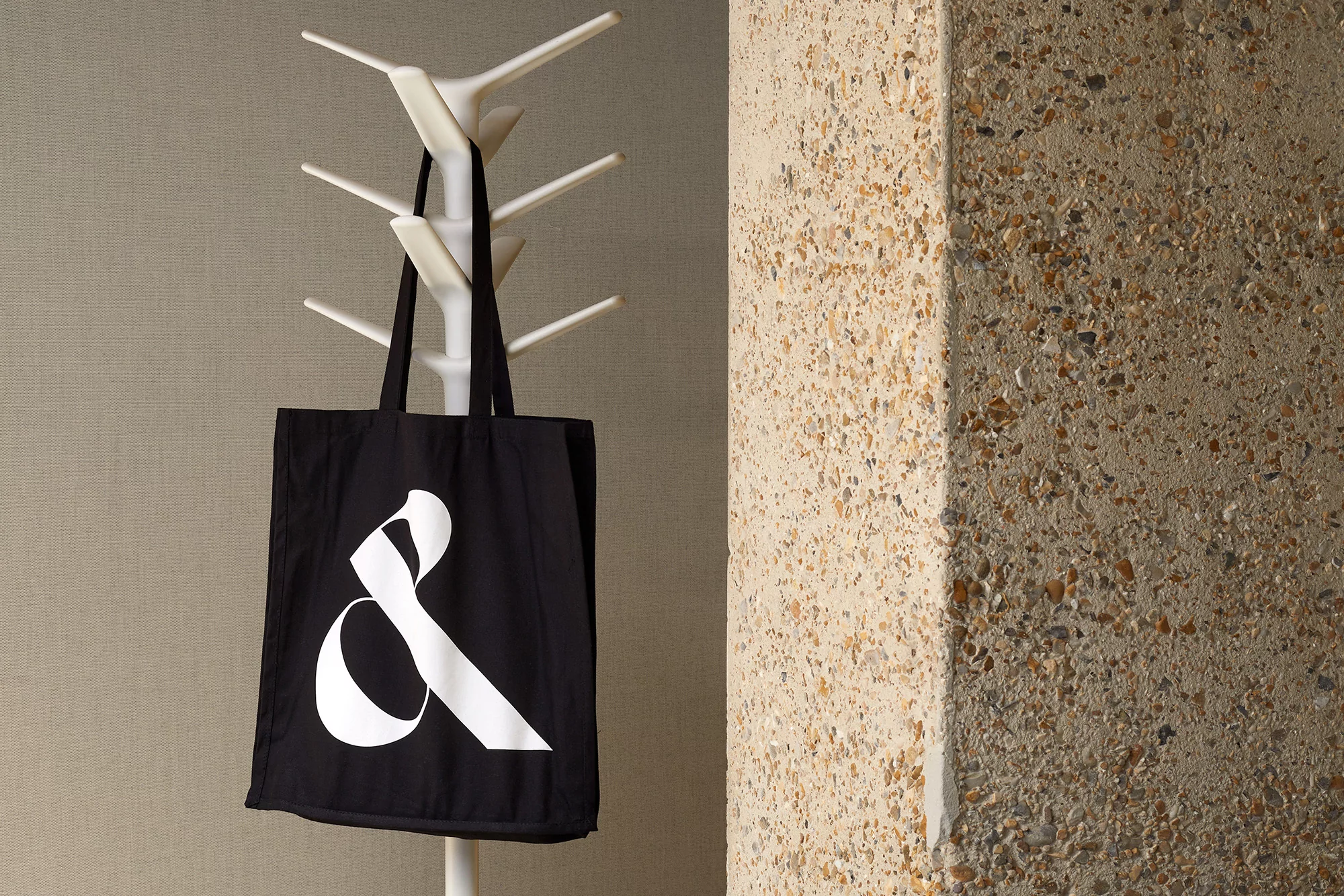 Black tote bag with white ampersand hangs on a white coat rack against a textured wall, showcasing modern design and interior decor.