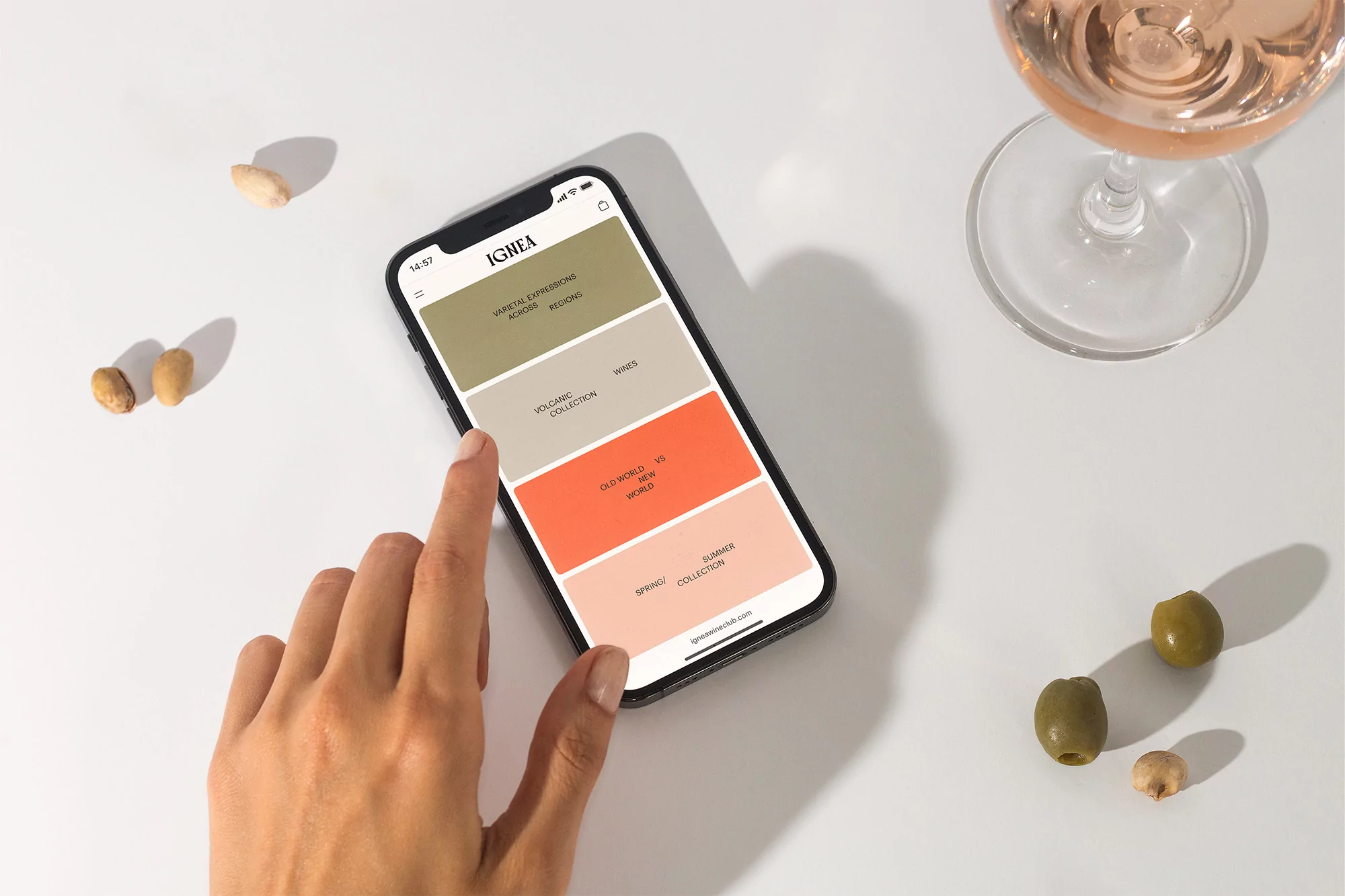 Hand interacts with a phone displaying a wine app, alongside a glass of rosé, olives, and pistachios on a bright surface.