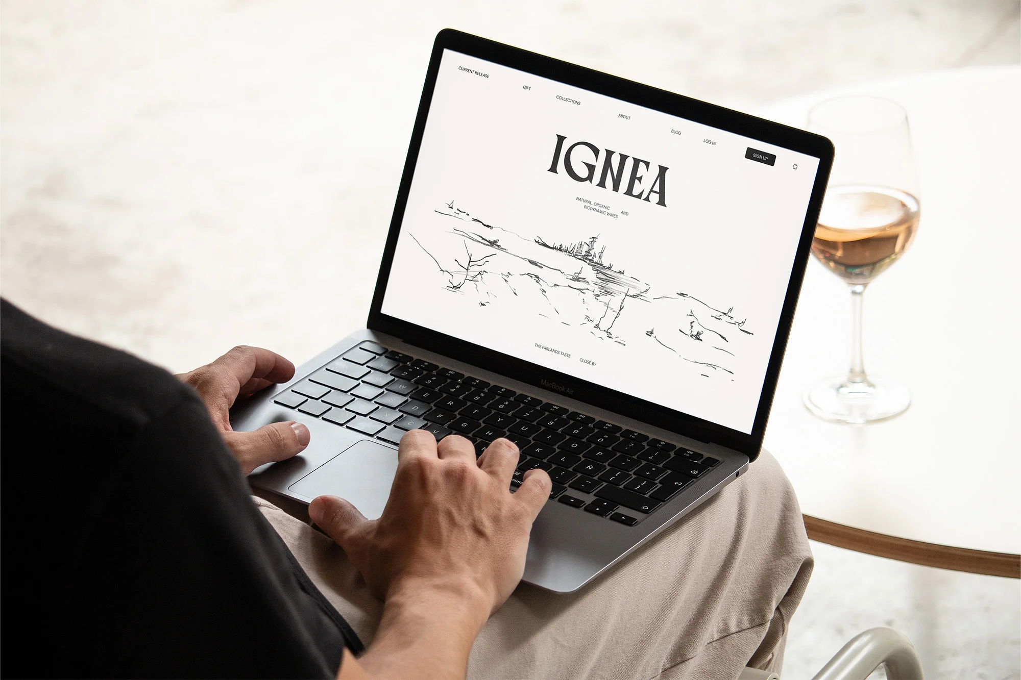 Man using a laptop displaying a website with a landscape drawing, with a glass of rose wine in the background on a white table.