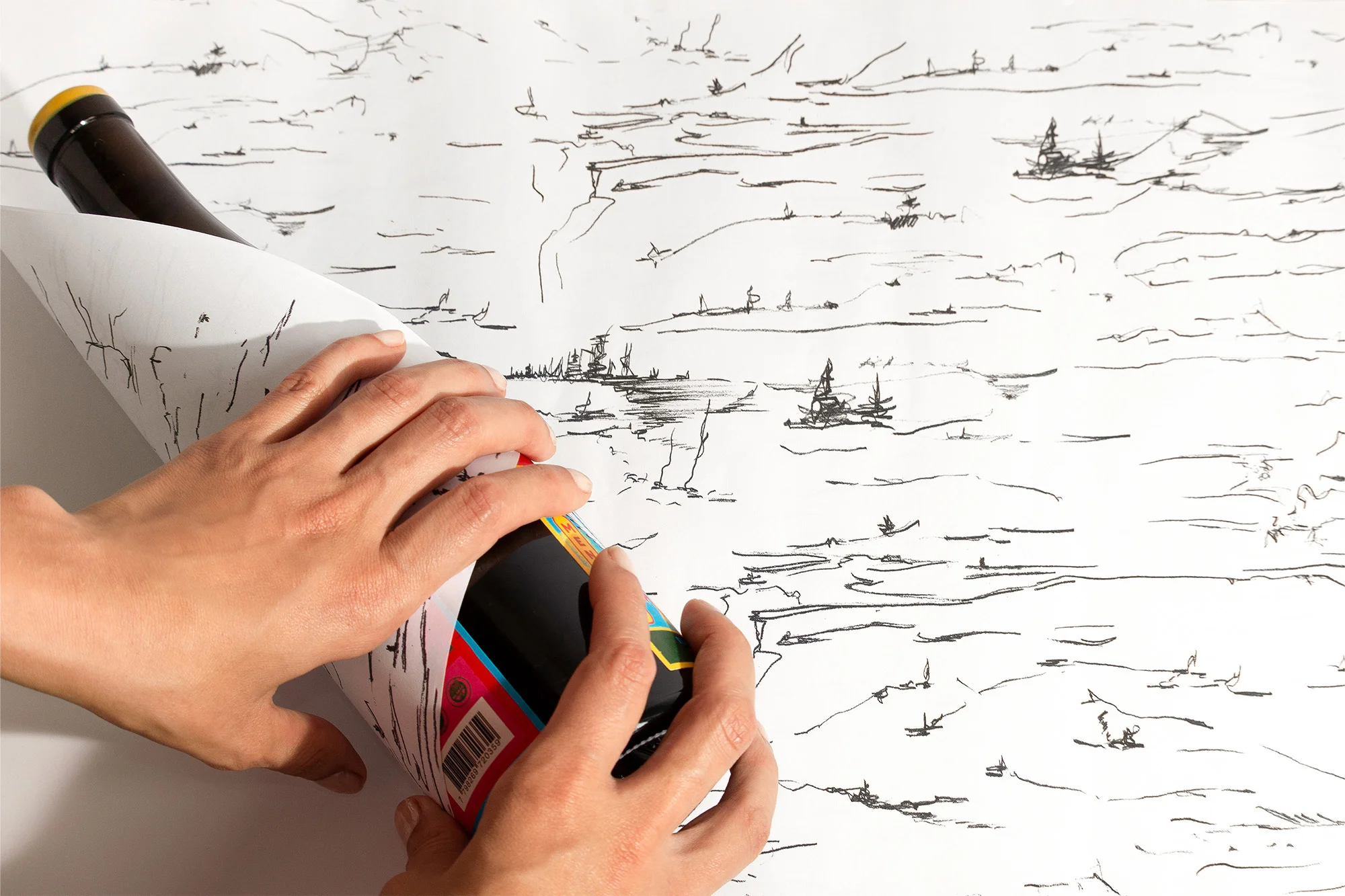 Close-up of hands wrapping a wine bottle with a hand-drawn landscape paper, set against a white background; unique presentation.