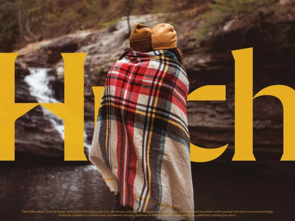 A couple wearing beanies and wrapped in a red and white plaid blanket are standing in front of a waterfall and brown cliffs.