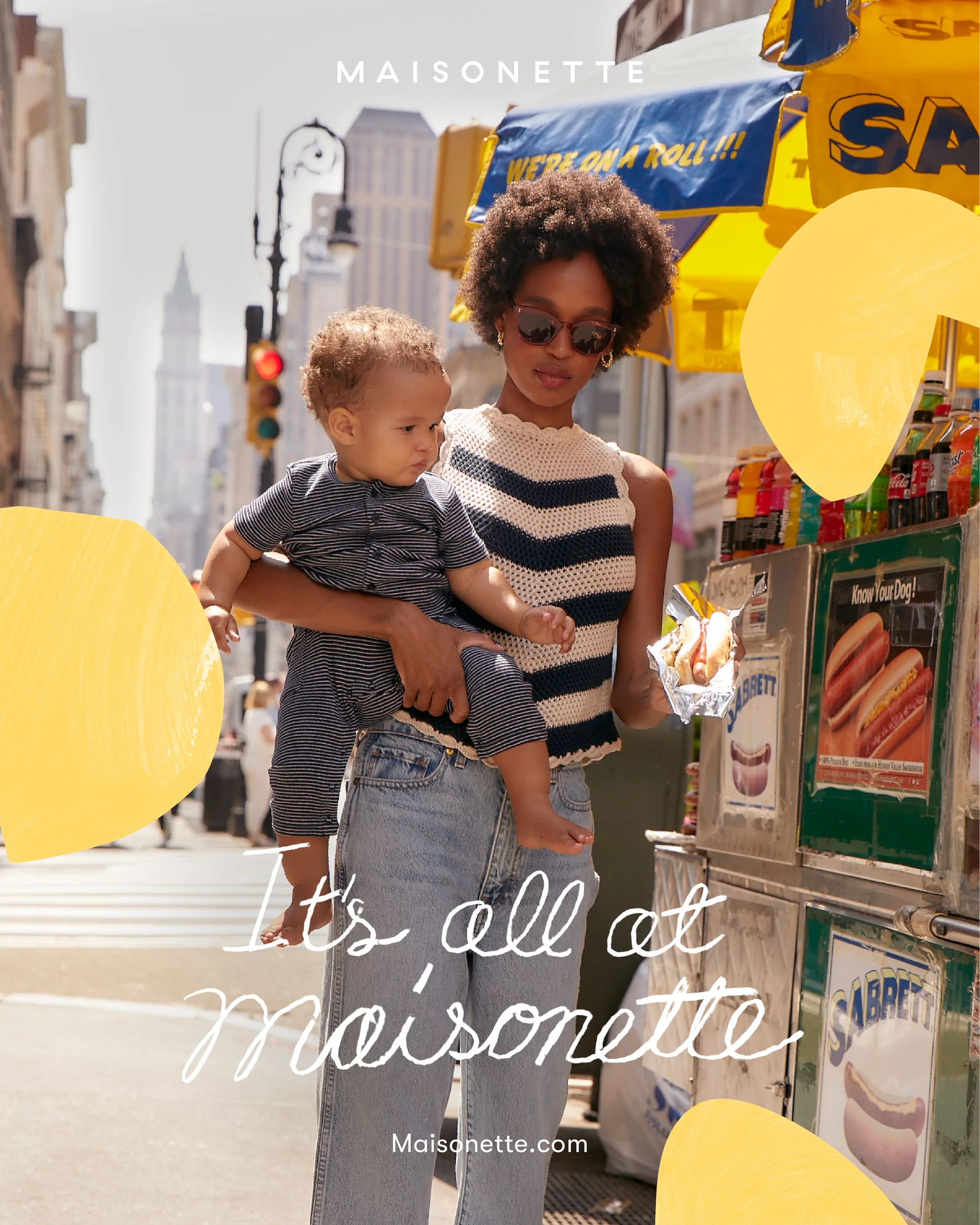 A young mother carries her toddler son in an urban setting, standing near a hot dog stand on a sunny day, with city buildings in the background.