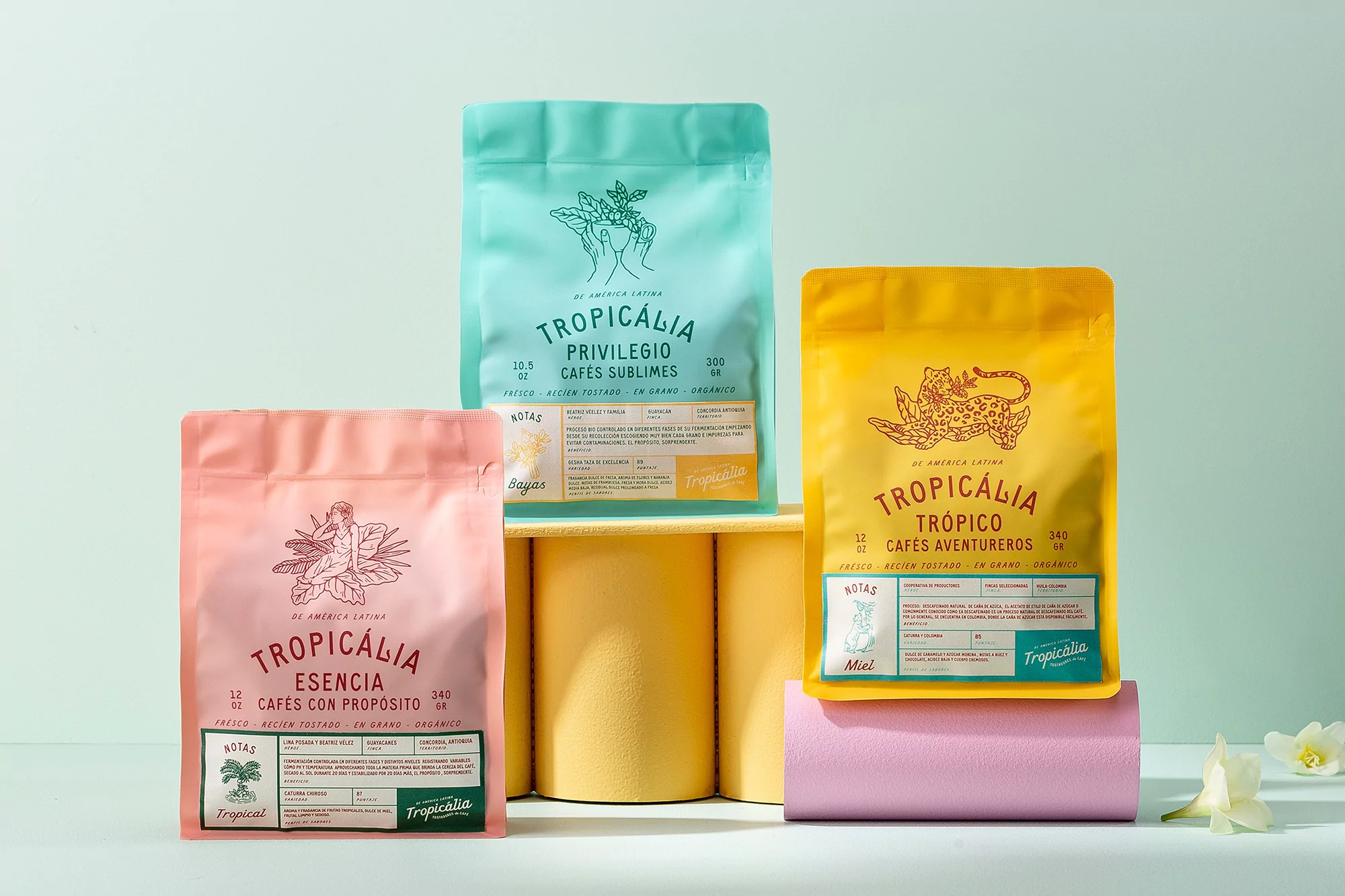 Three colorful Tropícalia coffee bags: a pink bag, a turquoise bag, and a yellow bag. The coffee bags are stacked on yellow and pink platforms.